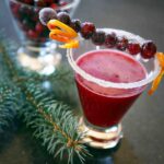 A cranberry margarita in a short glass with a skewer or sugared cranberries and orange zest across the top.