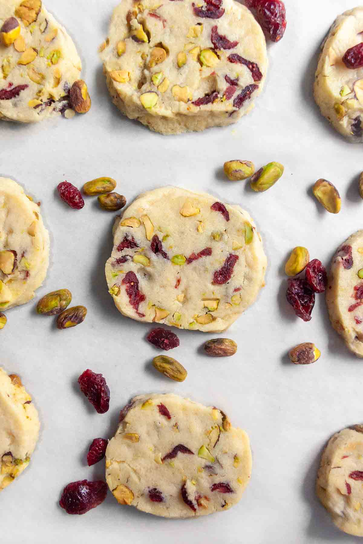 Nine cranberry pistachio cookies on a sheet of parchment with pistachios and dried cranberries scattered around them.