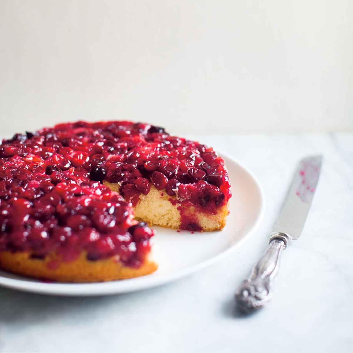 Cranberry upside-down cake with a large piece missing, on a white plate with a serving knife beside it.