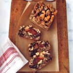 Dried fruit and nut cake in slices on a wooden cutting board with parchment paper, beside a red and white dish towel.