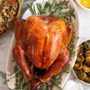 A whole roasted dry brined turkey on an oval platter on a bed of herbs with side dishes around it.