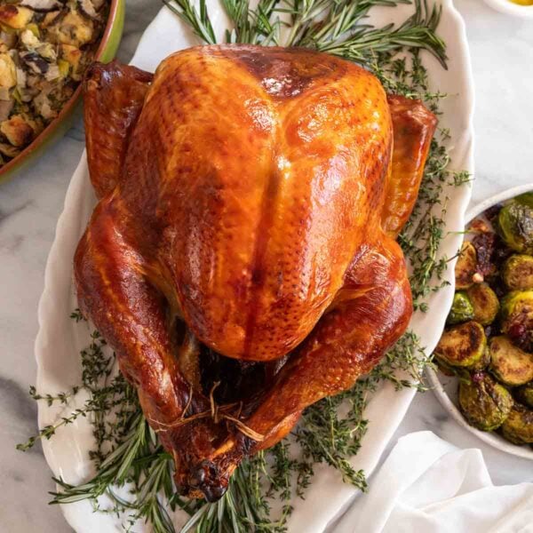 A whole roasted dry brined turkey on an oval platter on a bed of herbs with side dishes around it.