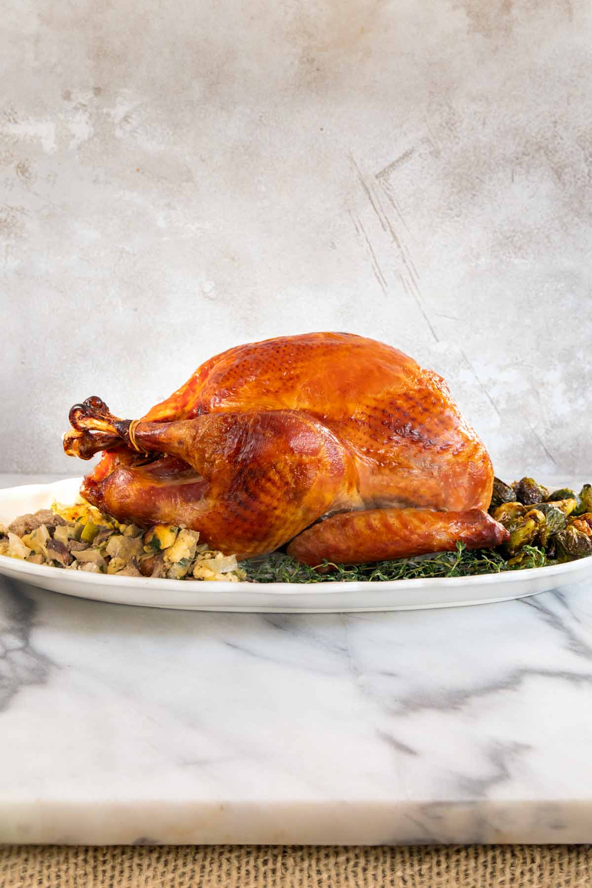A cooked dry brined turkey on a bed of herbs with stuffing and roasted Brussels sprouts around it.