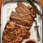 An enamel baking dish holding a sliced oven brisket topped with onions with jus on the side.