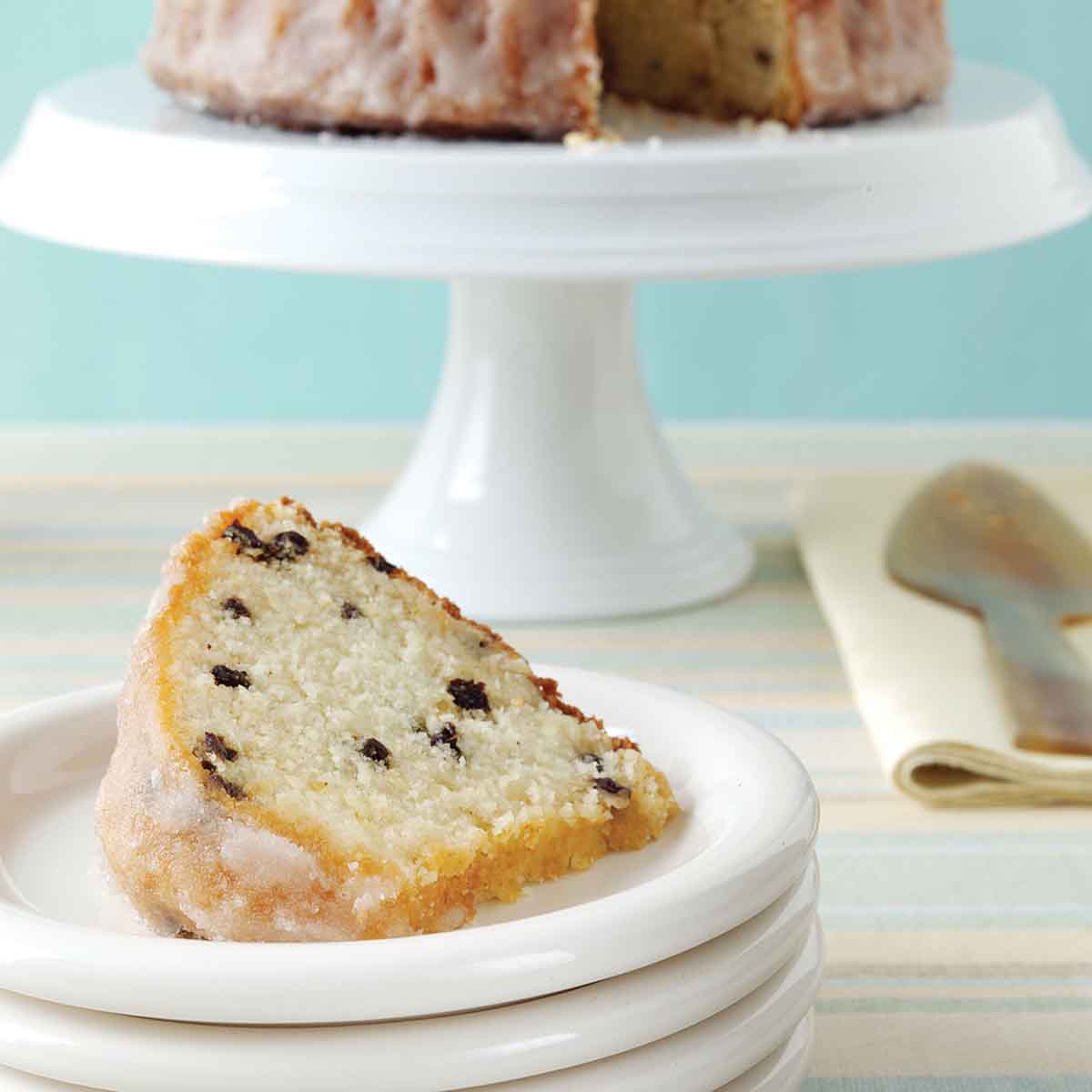 A plate with a slice of pound cake with currants, and a cake stand in the background with the remaining cake.