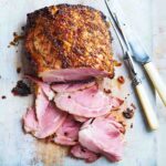 A glazed ham on a cutting board with several slices cut from the end and a carving fork and knife resting beside it.
