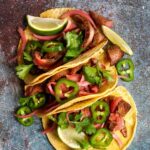 Sliced skirt steak on a wooden board with tortillas, lime wedges, cilantro, jalapeno, and pickled red onion.