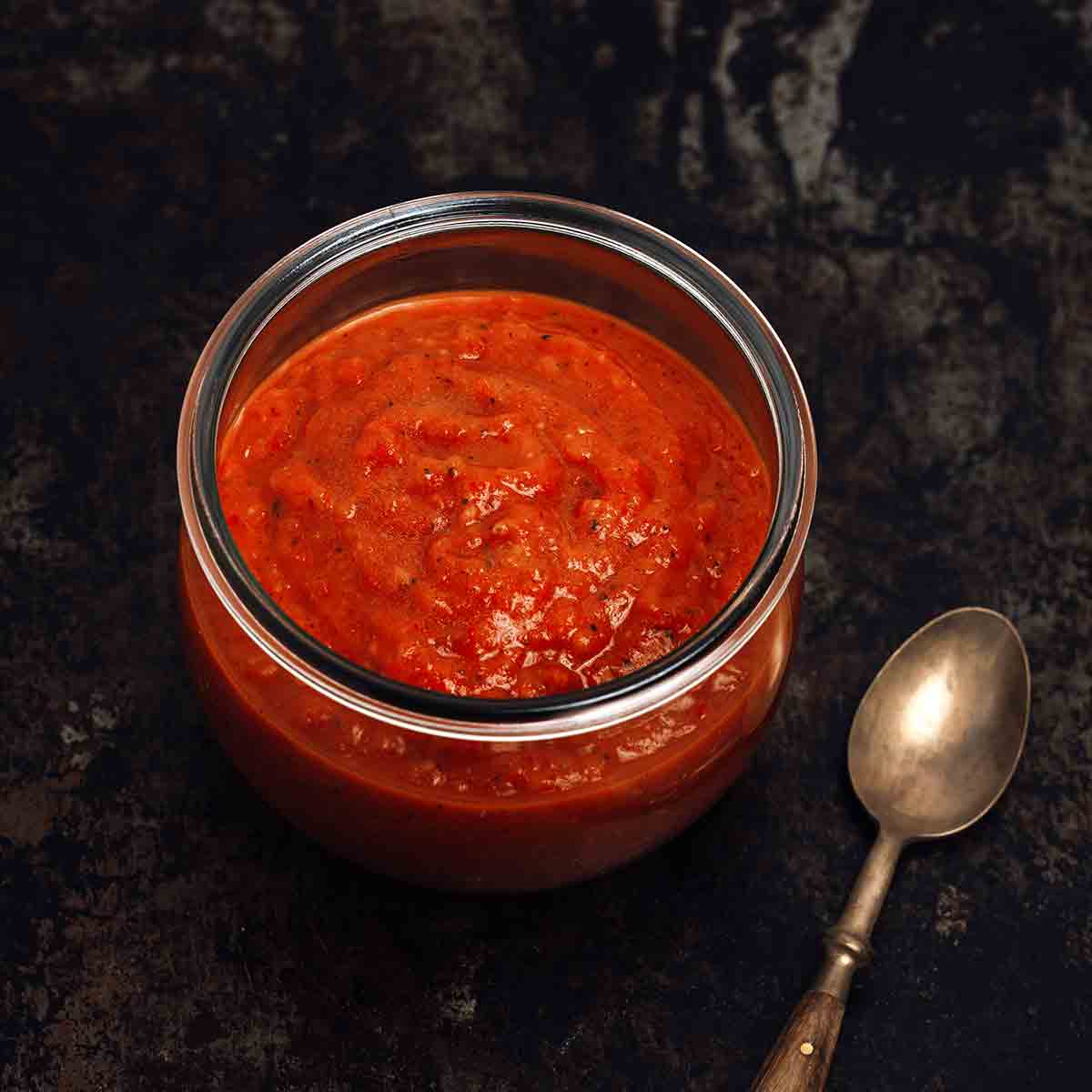 A jar of red homemade sriracha hot sauce on a black background. A spoon nearby.