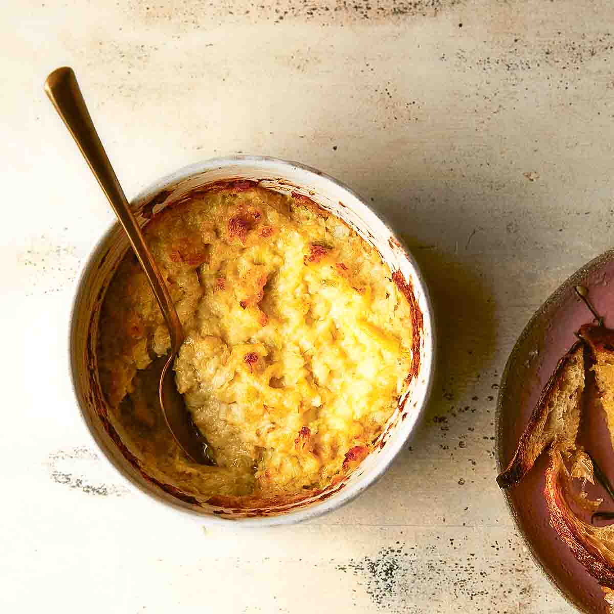 Hot artichoke dip in a white pottery bowl with a serving spoon, beside a bowl of toasted bread.