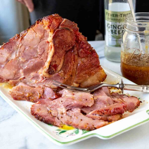 A partially carved ham on a platter with a bottle of ginger beer and a jar of sauce in the background.