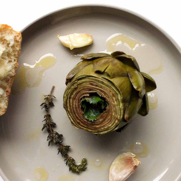 A braised artichoke on a round plate with a couple of garlic cloves, a thyme sprig, and piece of bread on the side.