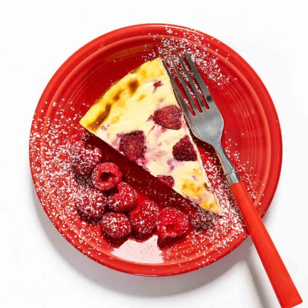 A slice of lemon raspberry cheesecake dusted with confectioners' sugar on a red plate with a fork and seven raspberries on the side.