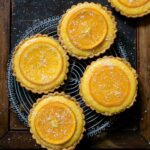 Five small tarts filled with orange curd and topped with candied orange slices on a cooling rack.