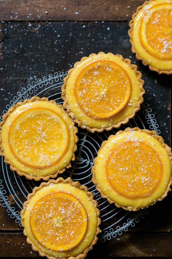 Five small tarts filled with orange curd and topped with candied orange slices on a cooling rack.