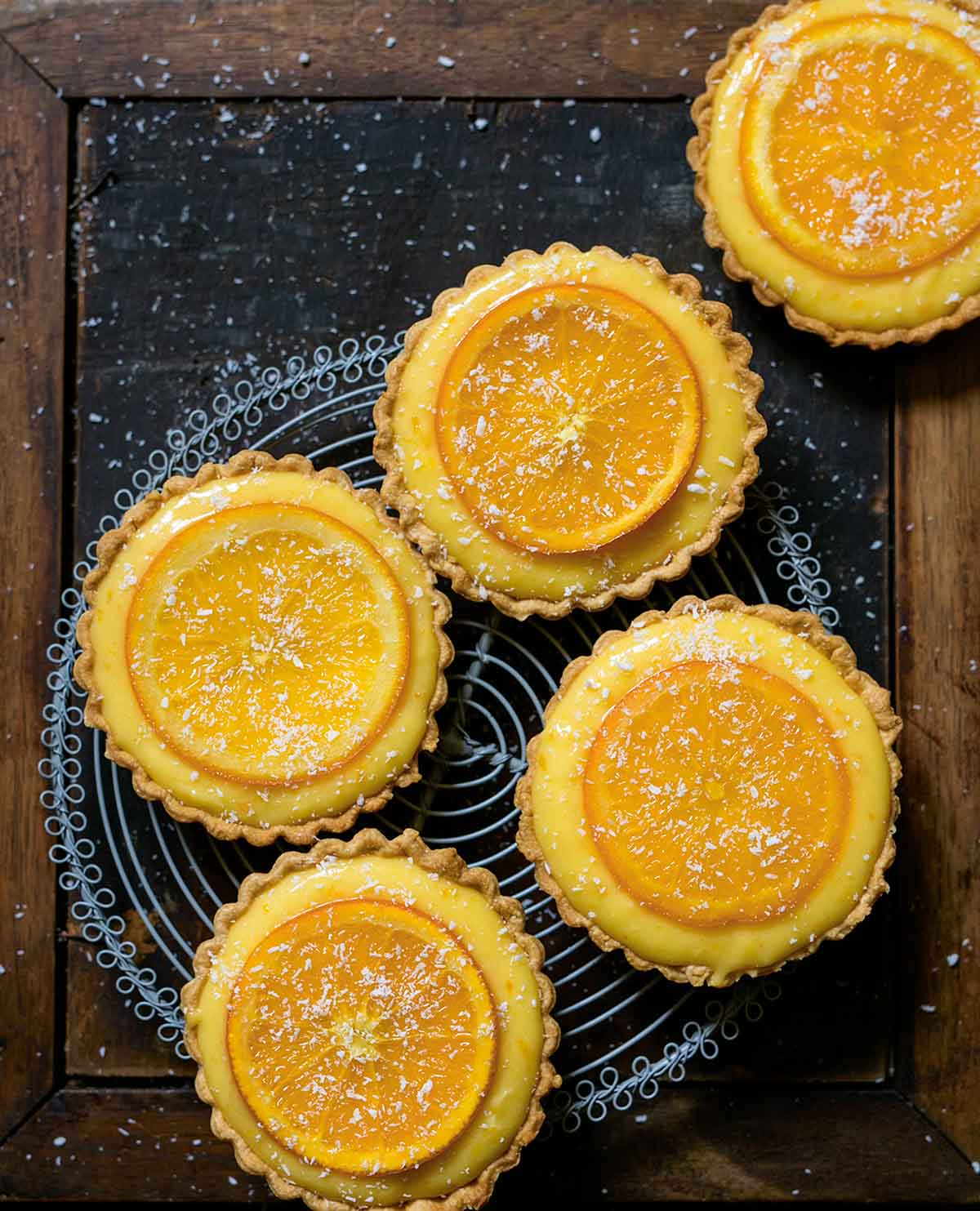Five small tarts filled with orange curd and topped with candied orange slices on a cooling rack.