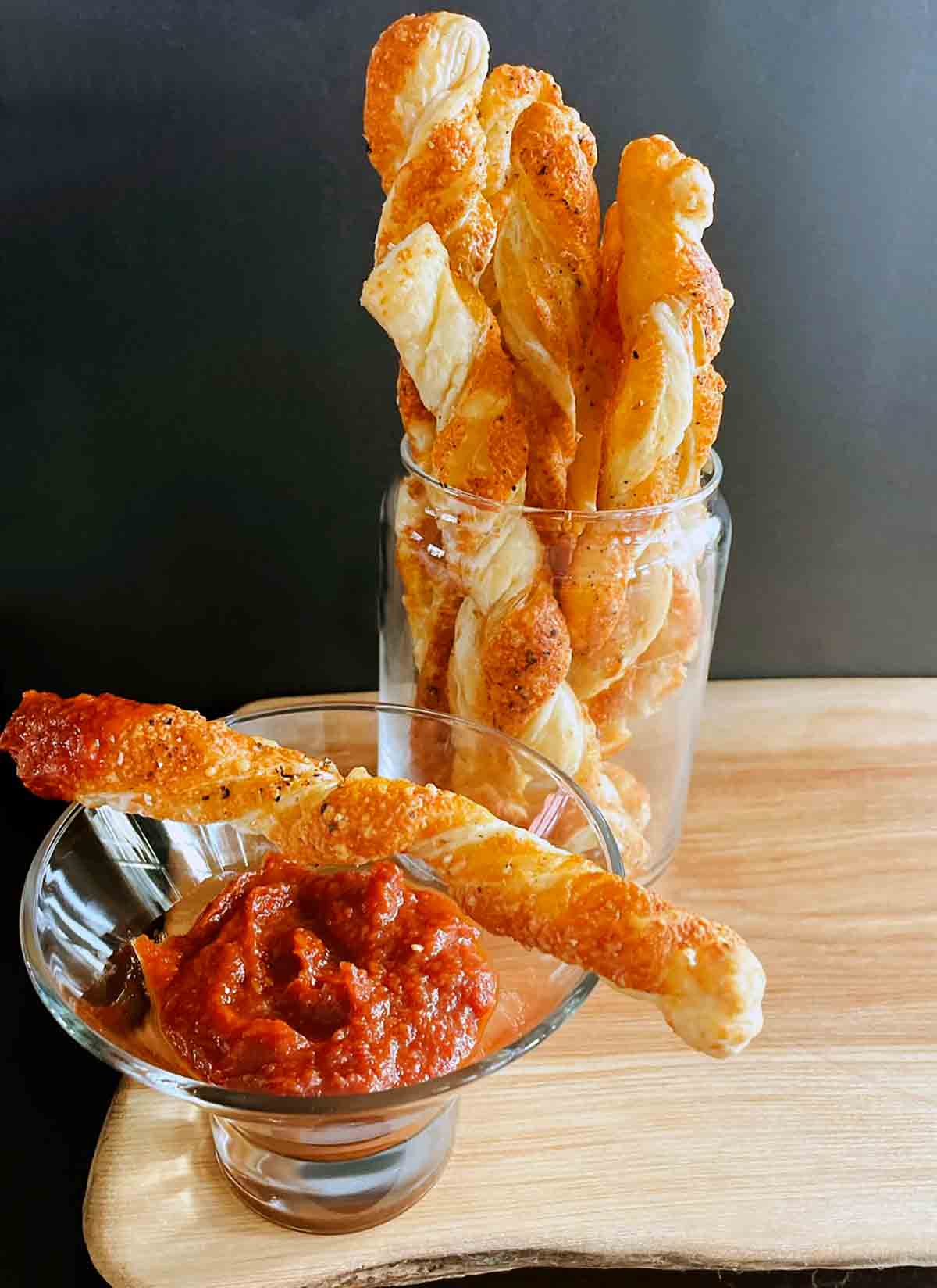 Peppery cheese straws arranged upright in a jar with one resting on top of a cocktail glass.