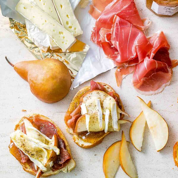 Two pieces of prosciutto, pear, and Brie toasts with slices of Brie, pear, prosciutto, and a jar of fig jam beside the toasts.