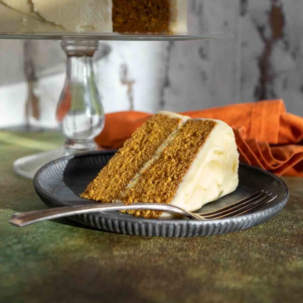 A slice of frosted pumpkin cake on a plate with the remaining cake on a stand in the background.
