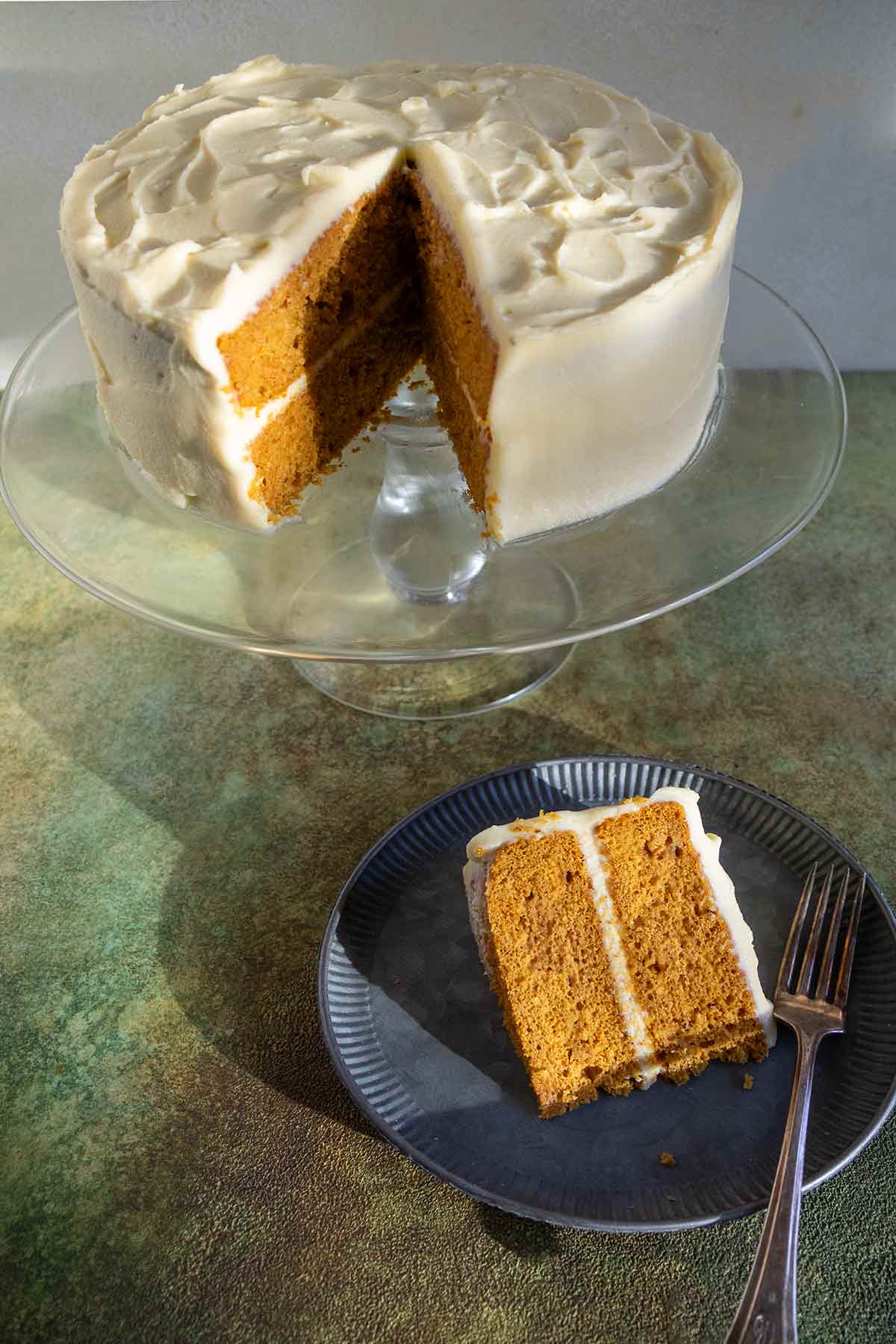 A slice of pumpkin cake with cream cheese frosting cut from a whole cake on a cake stand.