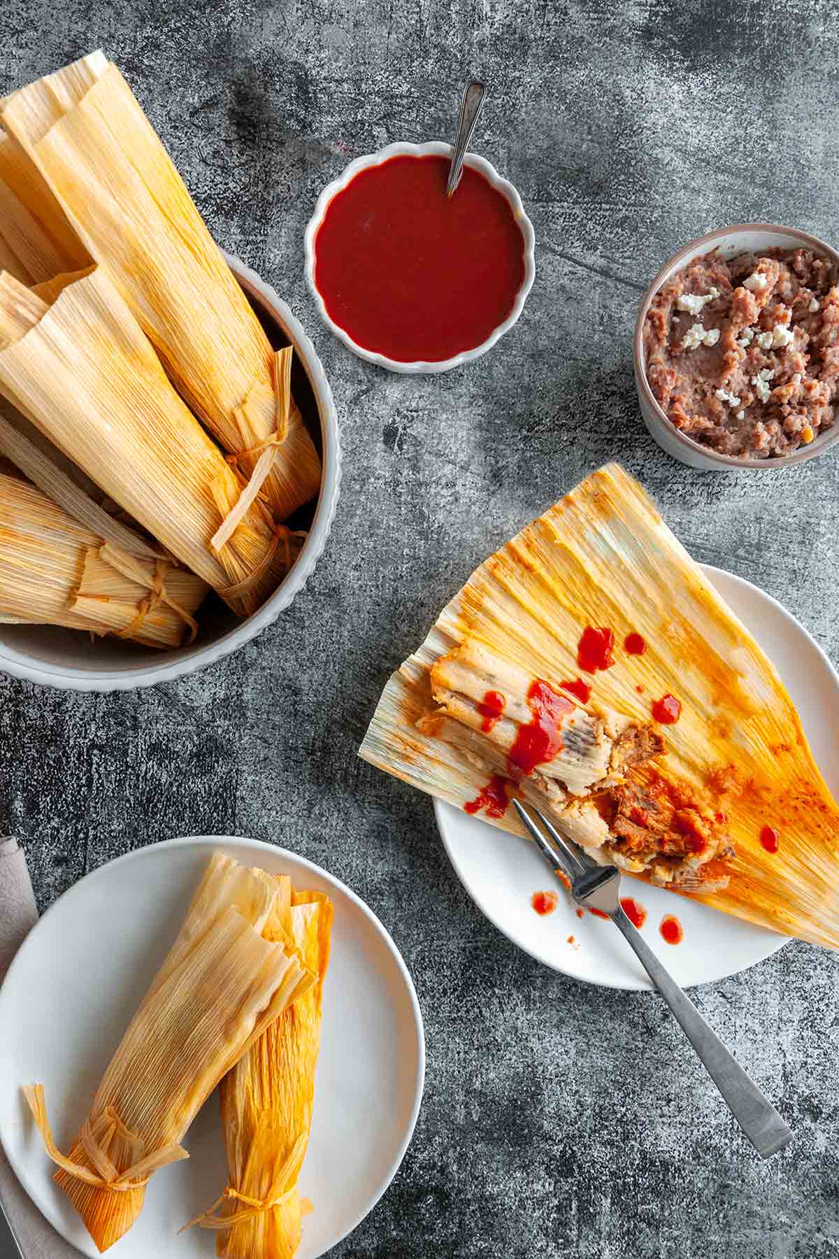 An open pork tamale, sprinkled with hot sauce with a fork resting on the side.