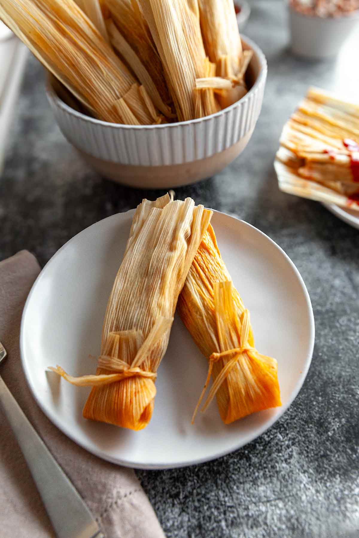 Two tamales on a white plate with a bowl of tamales in the background.