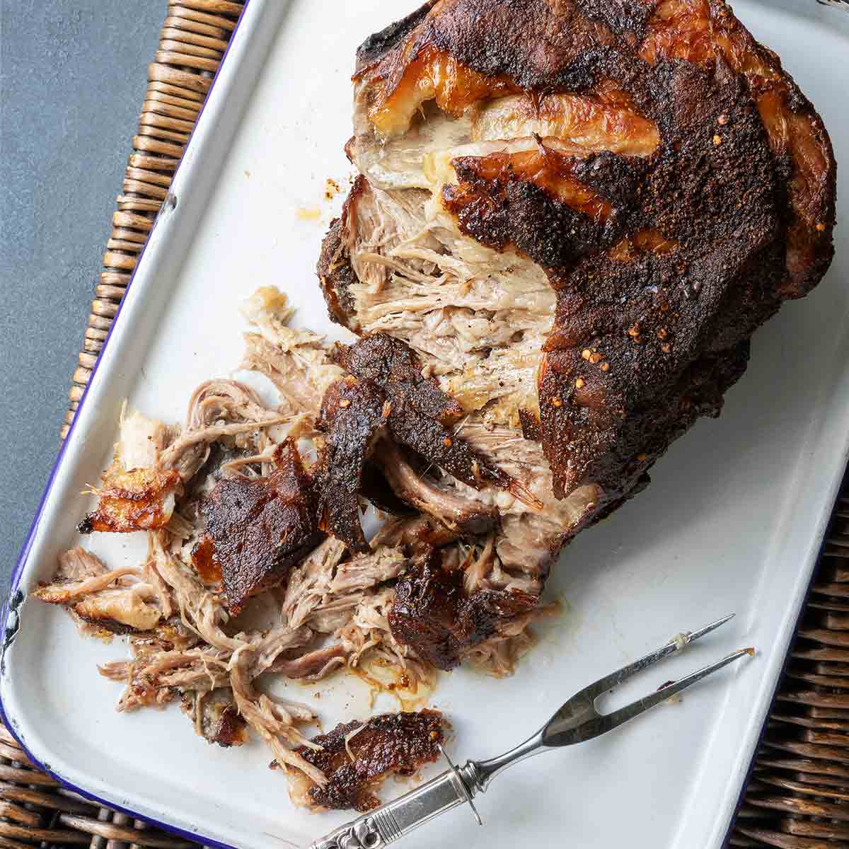 A partially shredded roast pork butt in a roasting pan with a fork nearby.