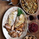 An oval platter with roasted and braised turkey, a dish of stuffing, a bowl of cranberry sauce, a bowl of chestnuts, two glasses of wine, and a small pot of gravy on a wooden table.