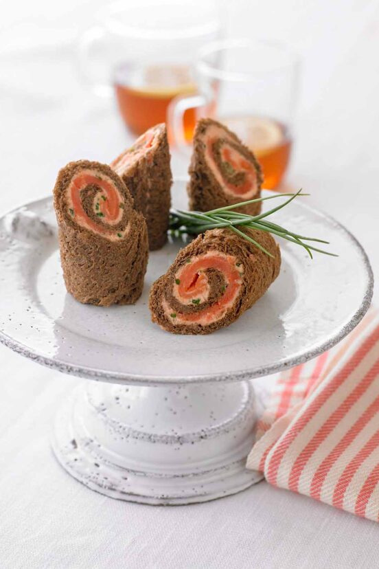 A white cake stand with four spirals of pumpernickel bread, salmon mousse, and smoked salmon.