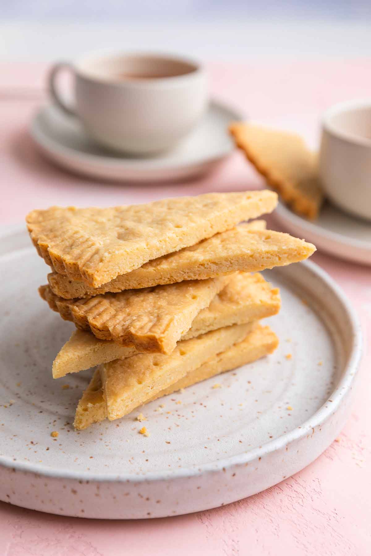 A stack of Scottish shortbread wedges on a plate with cups of tea nearby.