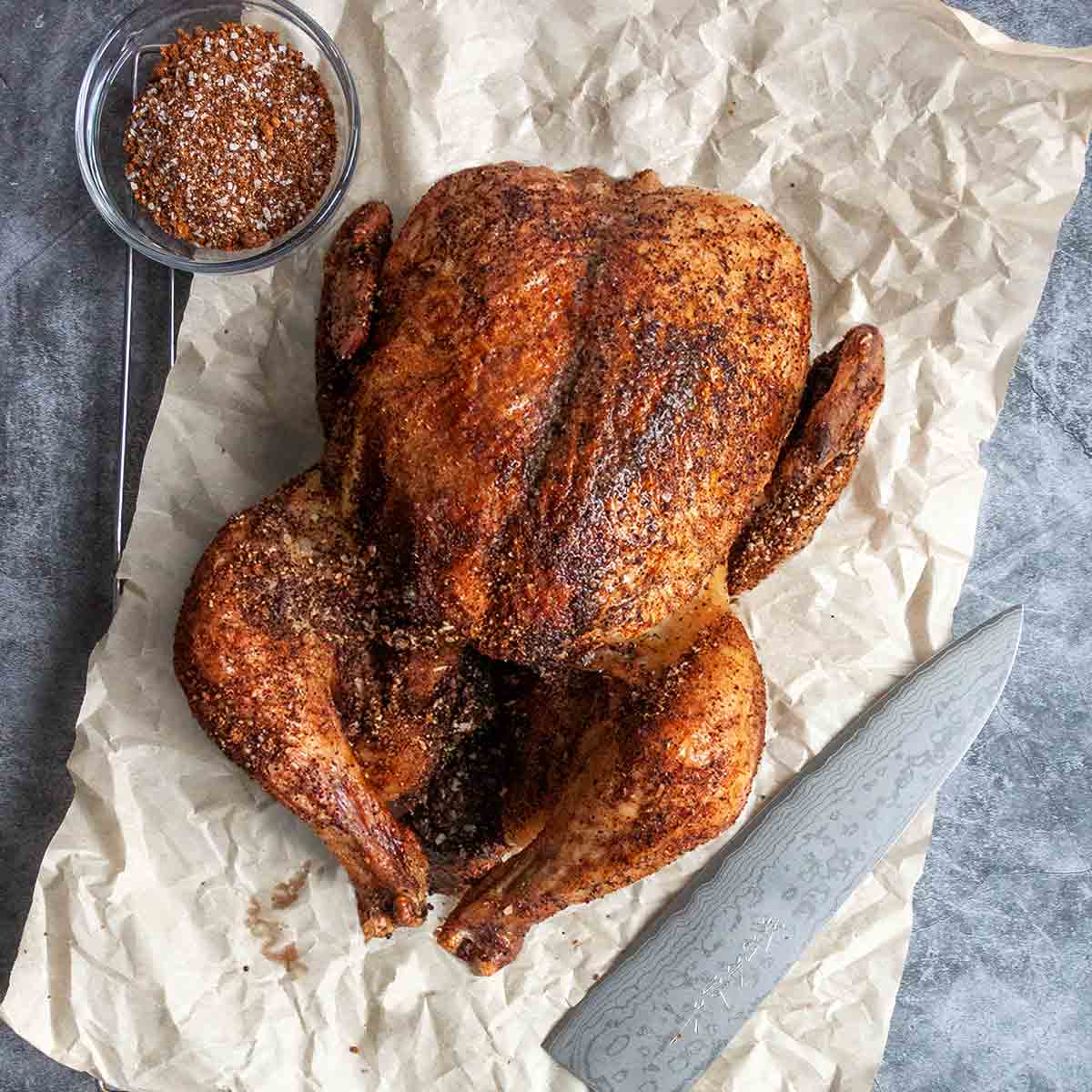 A smoked chicken on a piece of crumpled parchment paper with a knife and a bowl of spice rub on the side.