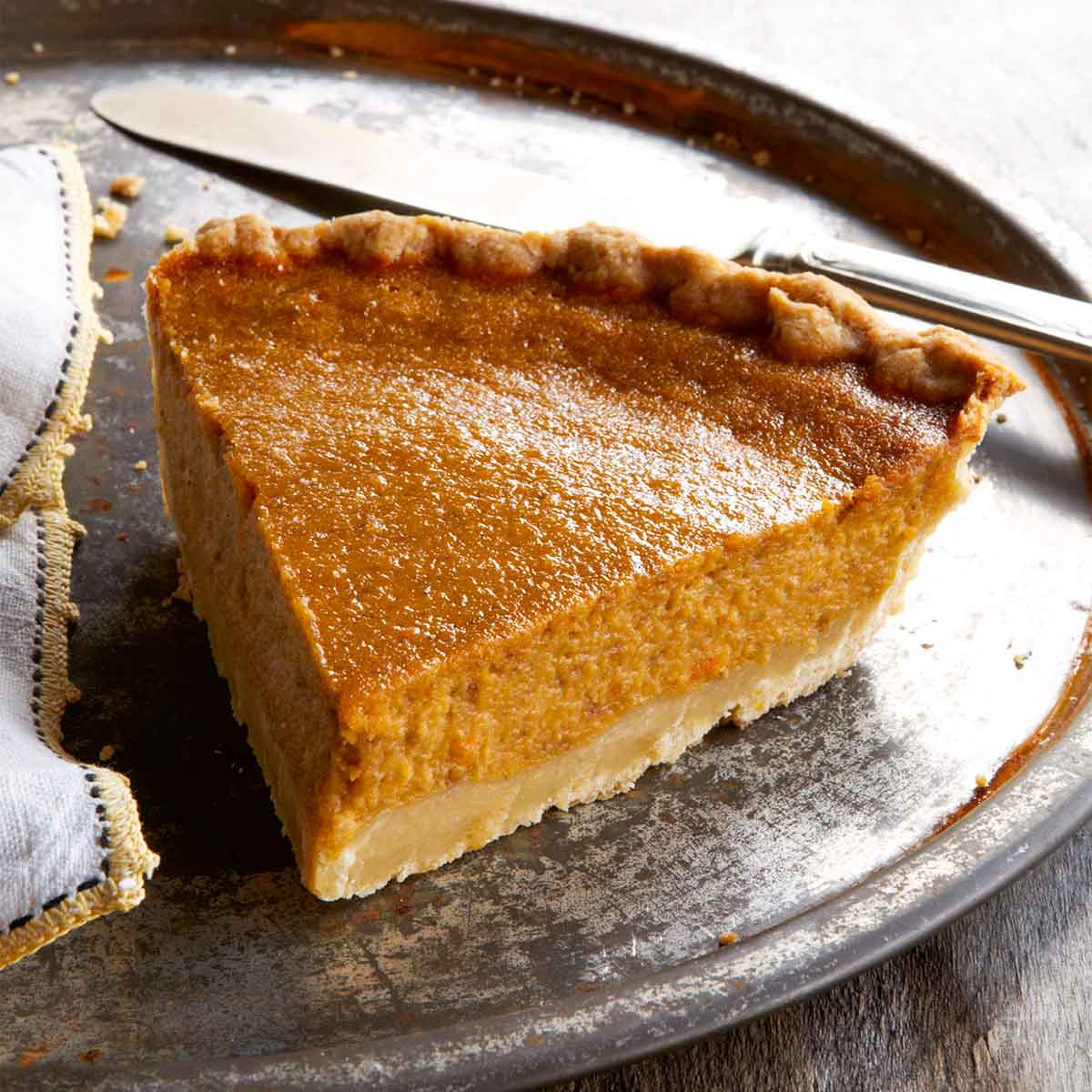 A slice of sweet potato pie on a large metal plate with a napkin and a knife.