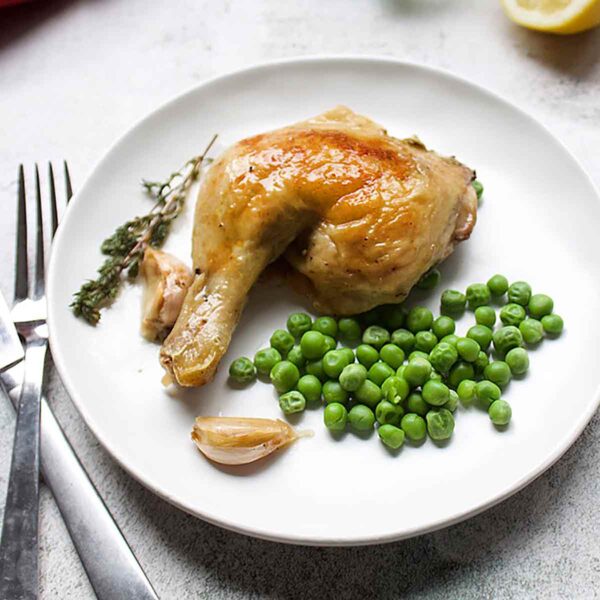 A roast chicken leg on a white plate with peas, thyme, and garlic.
