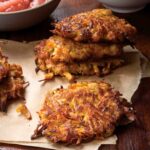 Several sweet potato and apple latkes on a paper towel with a bowl of applesauce in the background.