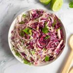 A bowl of apple coleslaw with wooden serving spoons, salt, two lime halves, and some cilantro on the side.