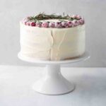 A whole frosted white Christmas cake topped with sugared cranberries and rosemary sprigs on a white cake stand.