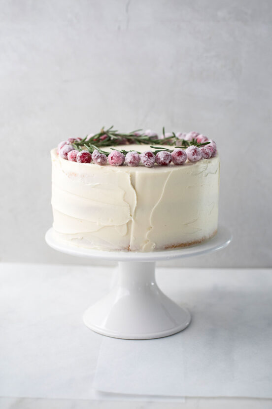 A whole frosted white Christmas cake topped with sugared cranberries and rosemary sprigs on a white cake stand.