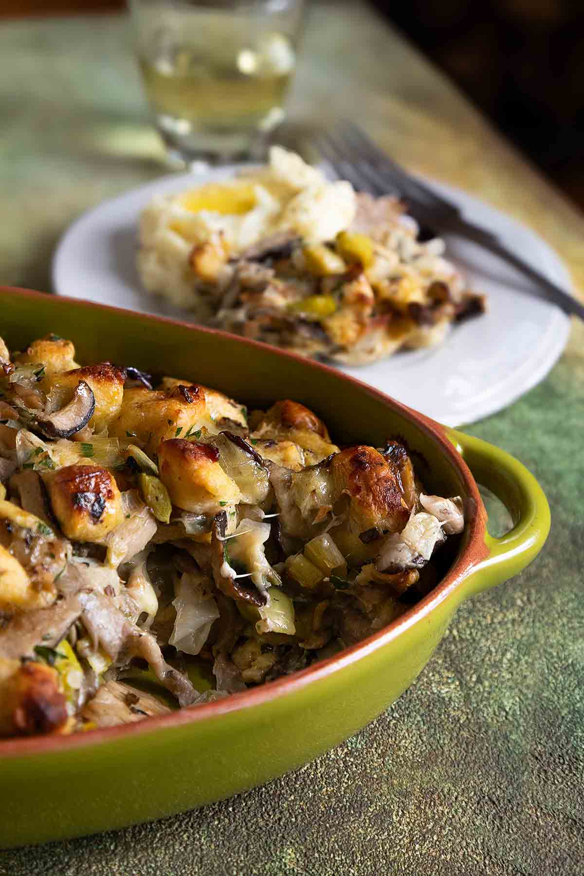 An oval casserole dish filled with wild mushroom stuffing and a plate of stuffing and mashed potatoes next to it.