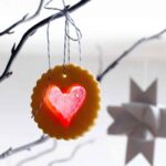 A scalloped round sugar cookie with a heart-shaped windowpane cutout hanging from a tree branch.