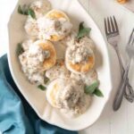 A serving platter with four cat head biscuits and gravy and sage leaves to garnish.