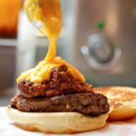 A chili cheddar burger on a bun half with cheese sauce being poured over it.