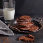 A metal tray with a pile of chocolate espresso shortbread cookies on it. Nearby, a glass of milk.