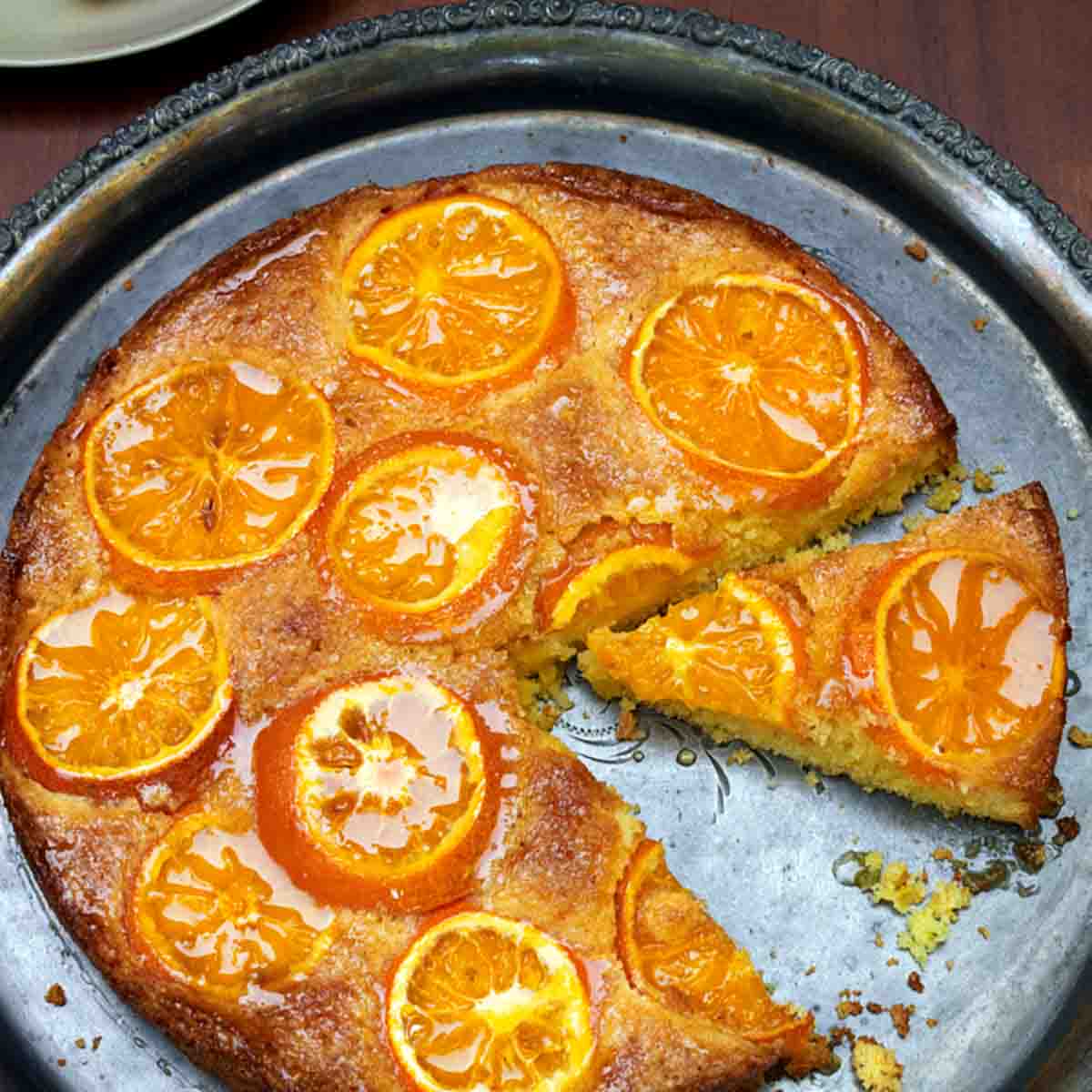 A clementine cake on a round silver platter with one slice on a plate beside it.