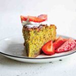 A slice of orange, olive oil, and poppyseed cake with sliced strawberries on the side on a white plate