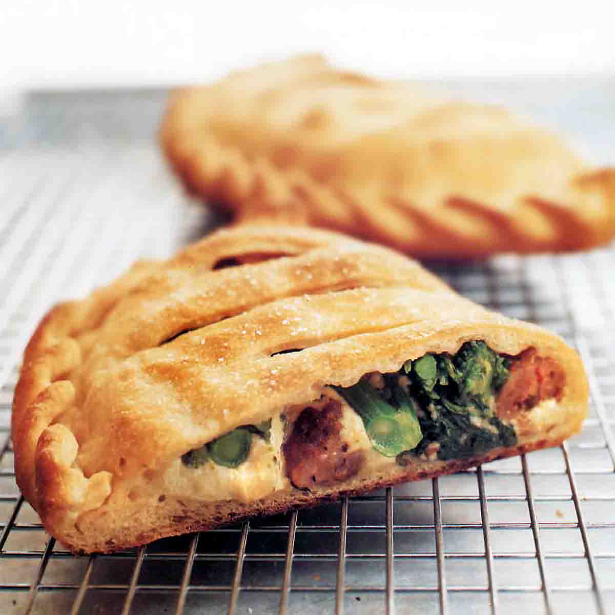Two ricotta calzones with sausage and broccoli rabe, one cut in half, the other whole, on a wire rack.