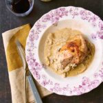 A floral-patterned plate topped with a piece of roast chicken with onion gravy around it and a fork, knife, and napkin on the side.