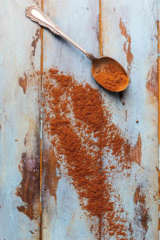 A spoonful of Shawarma spice blend on a wooden surface with some of the spice mix scattered nearby.