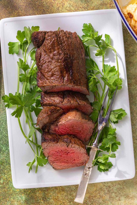A sliced beef tenderloin on a white platter with sprigs of parsley.