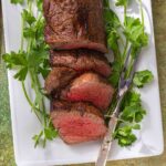 A sliced beef tenderloin on a white platter with sprigs of parsley.