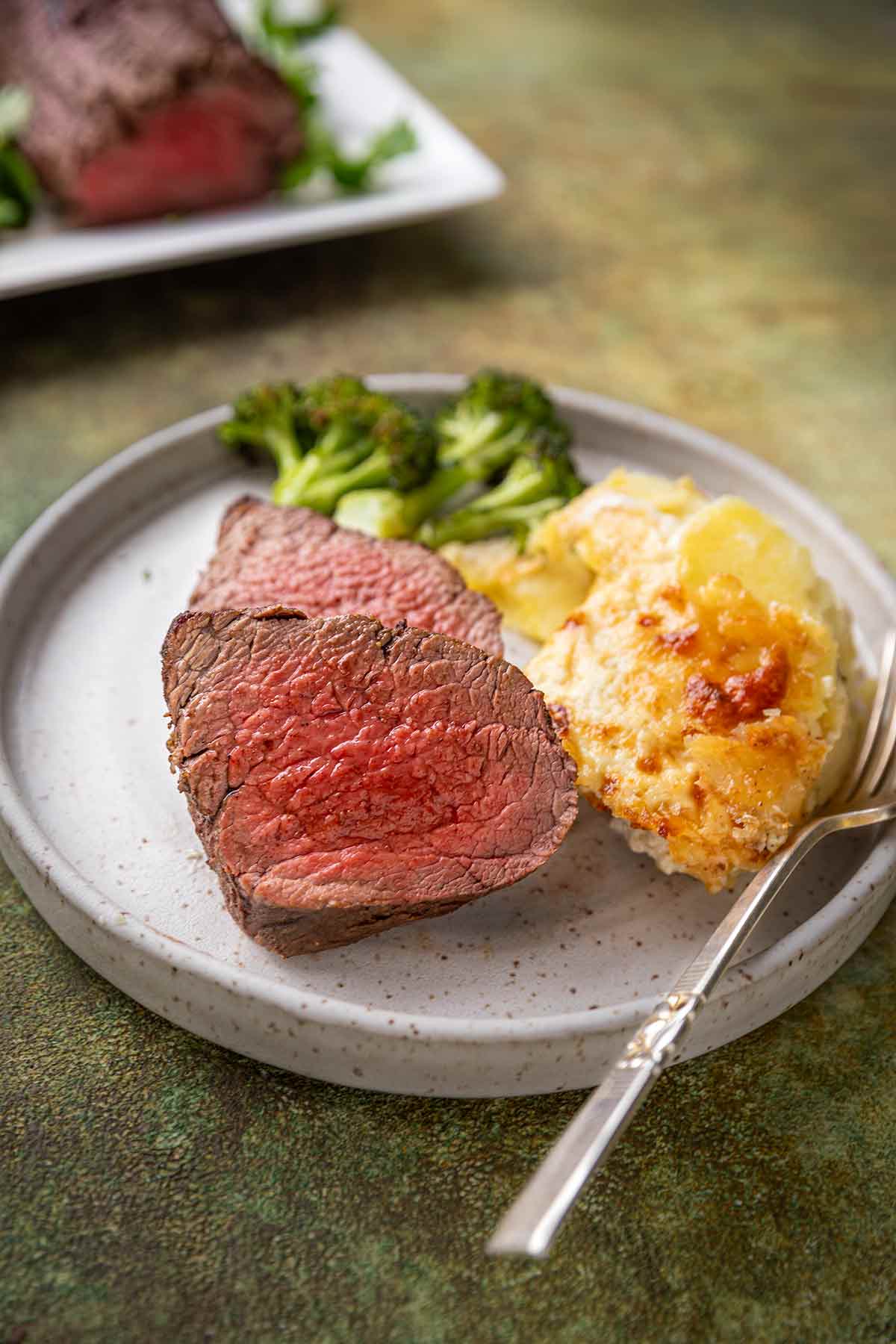 Slices of spiced beef tenderloin roast on a plate with cheesy potatoes and roasted broccoli.