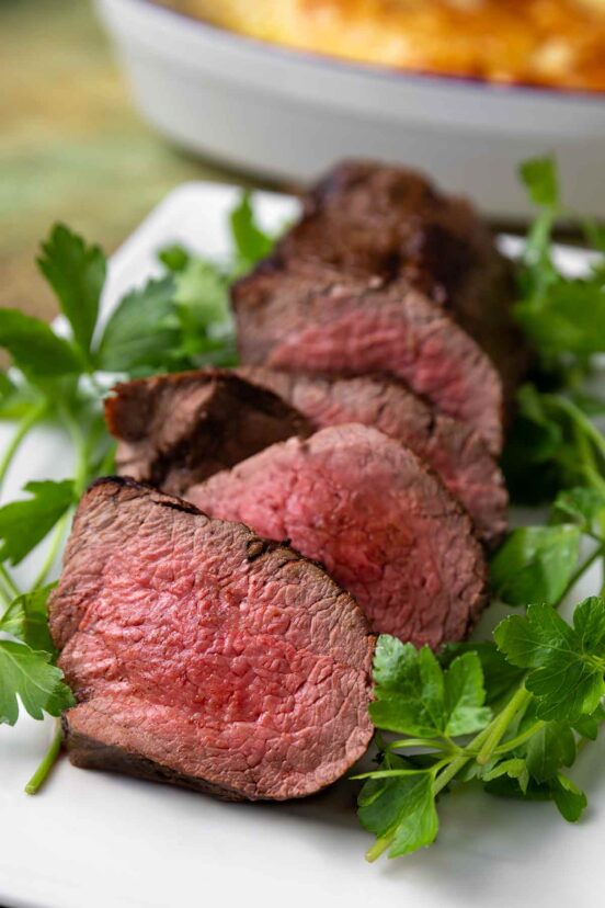 A sliced beef tenderloin on a white platter with sprigs of parsley.